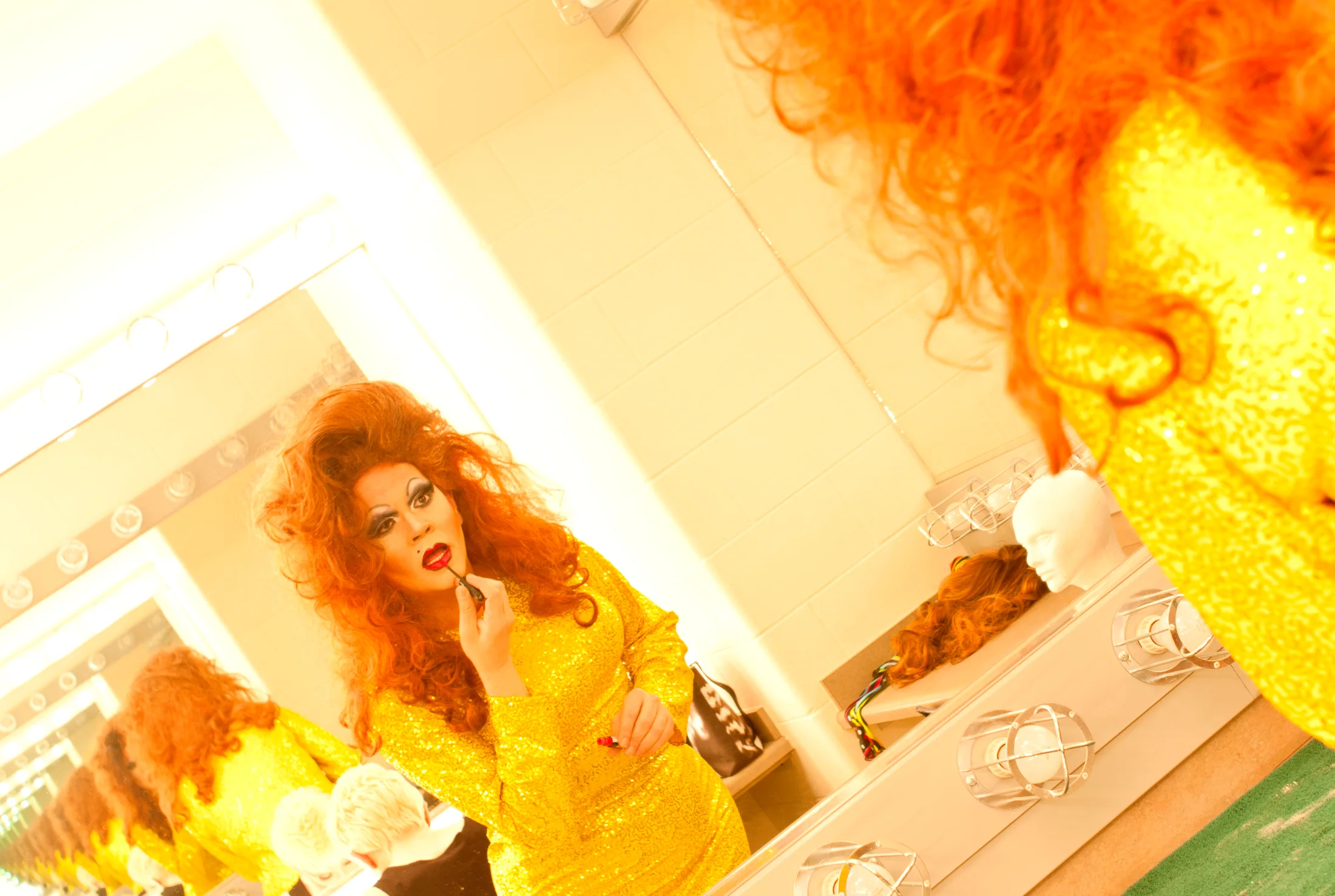A drag performer applies makeup in a warm backstage mirror scene.