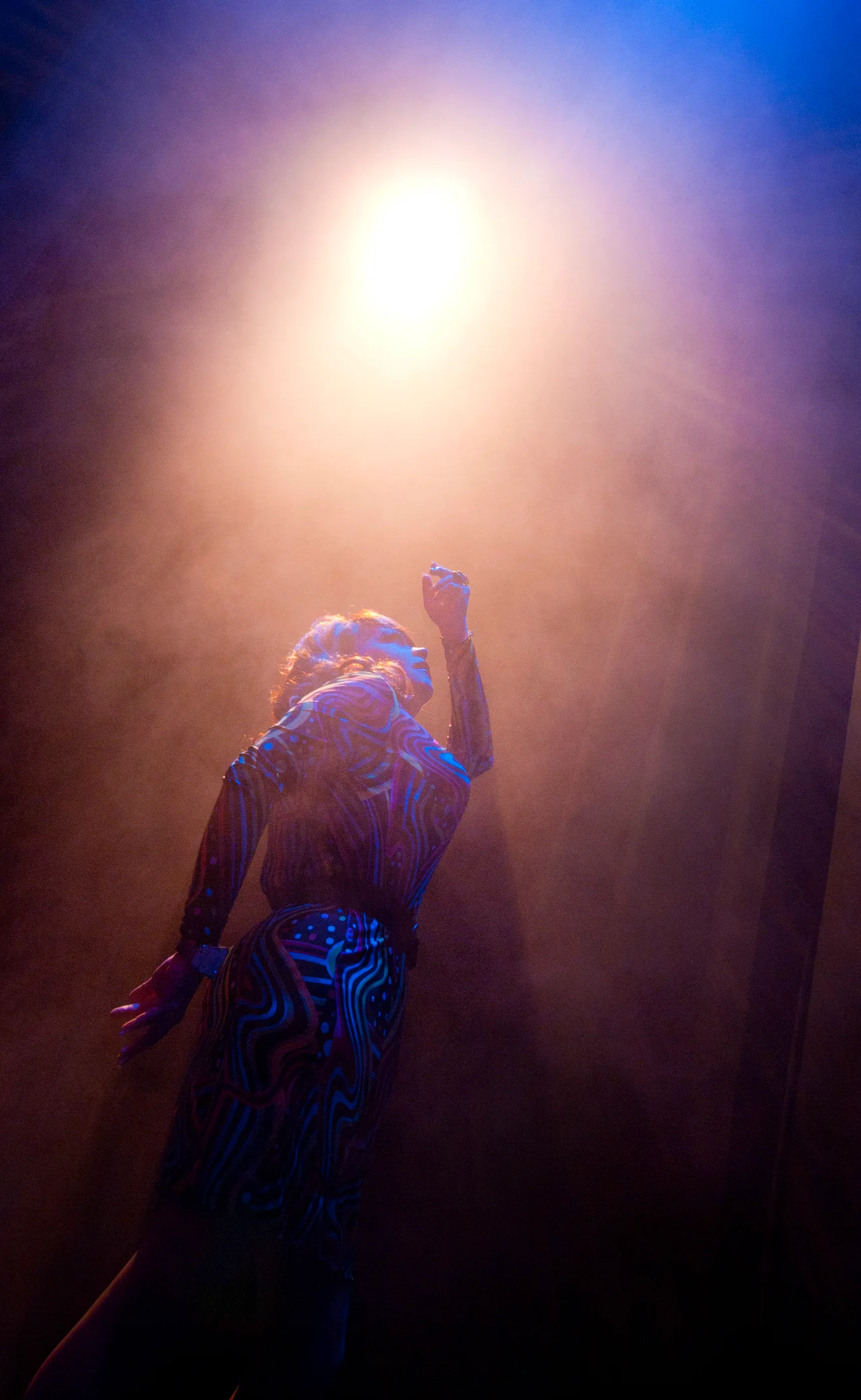 A drag performer stands in smoke beneath a bright overhead stage light.
