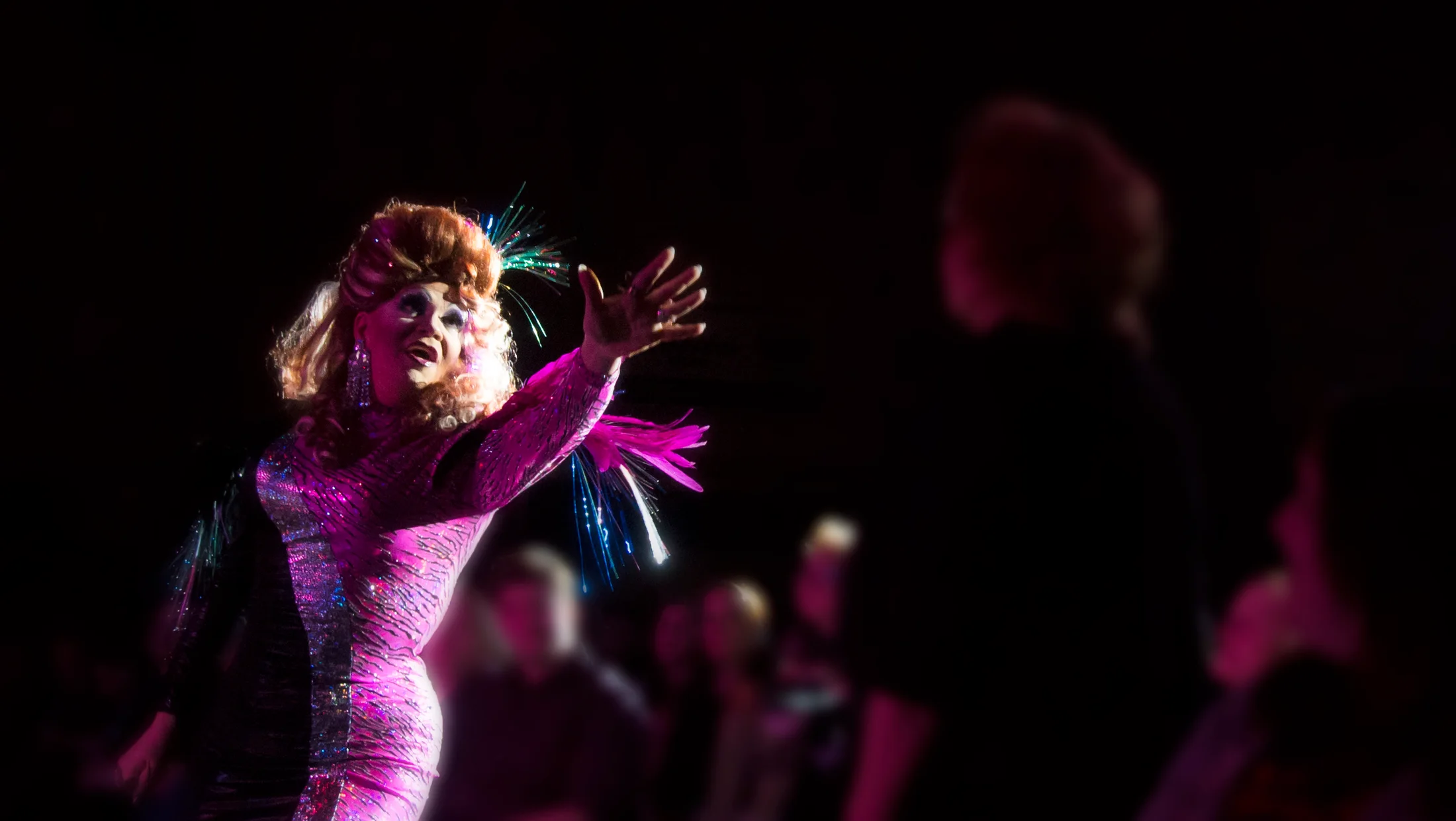 A drag performer reaches toward the crowd under magenta stage light.