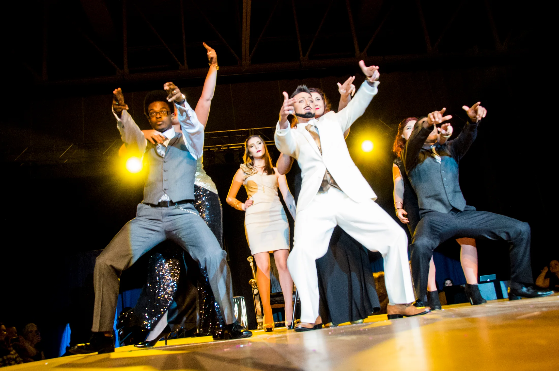 A group of performers points toward the audience under warm yellow stage light.