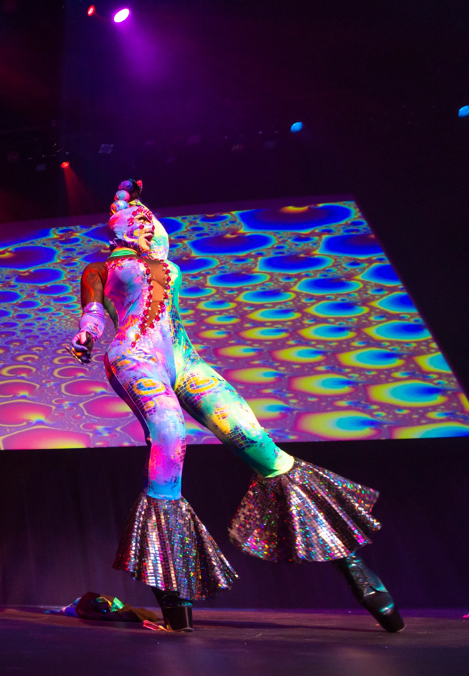 A drag performer in a colorful costume performs against projected stage light.