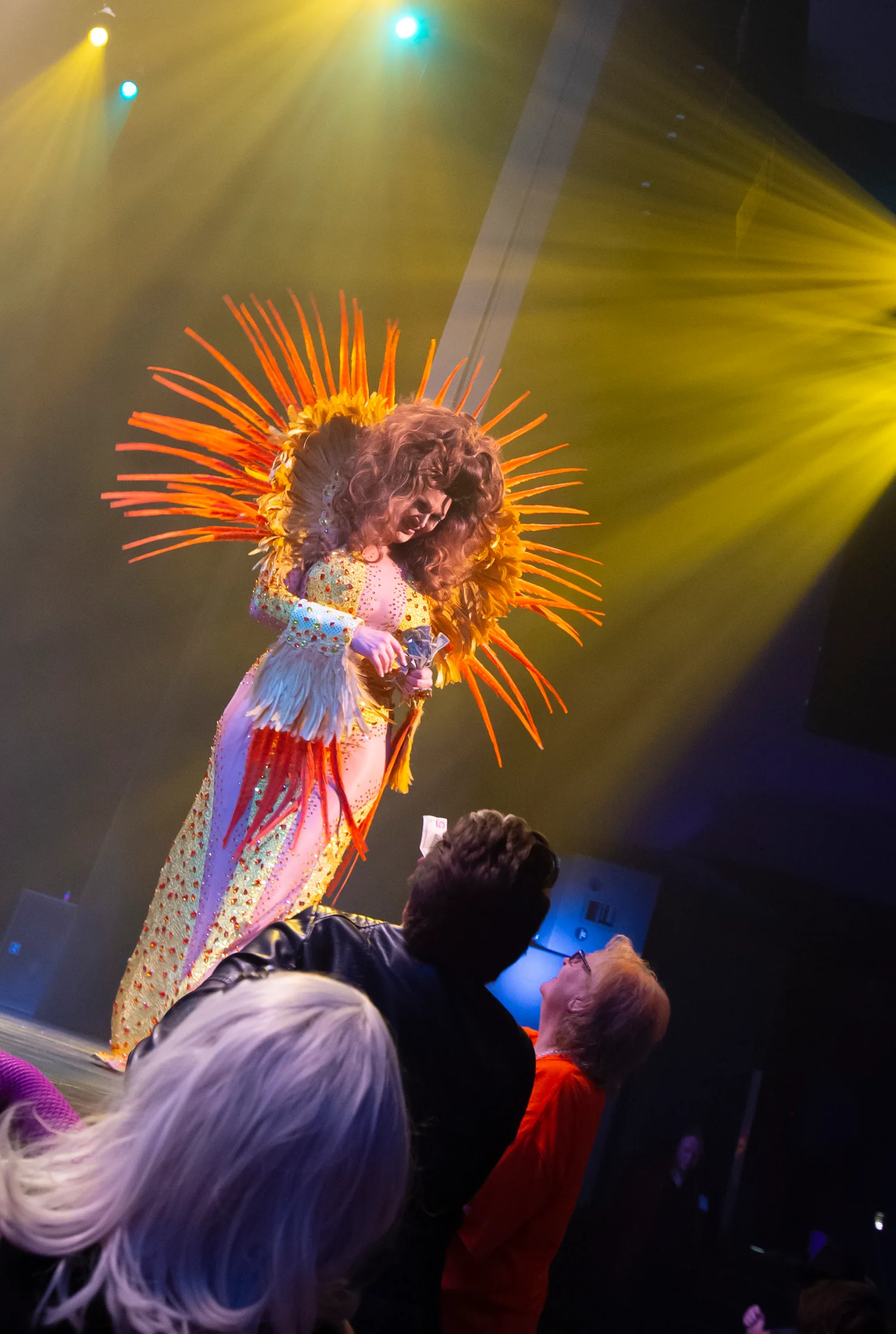 A drag performer in feathered stagewear receives a tip from an audience member.