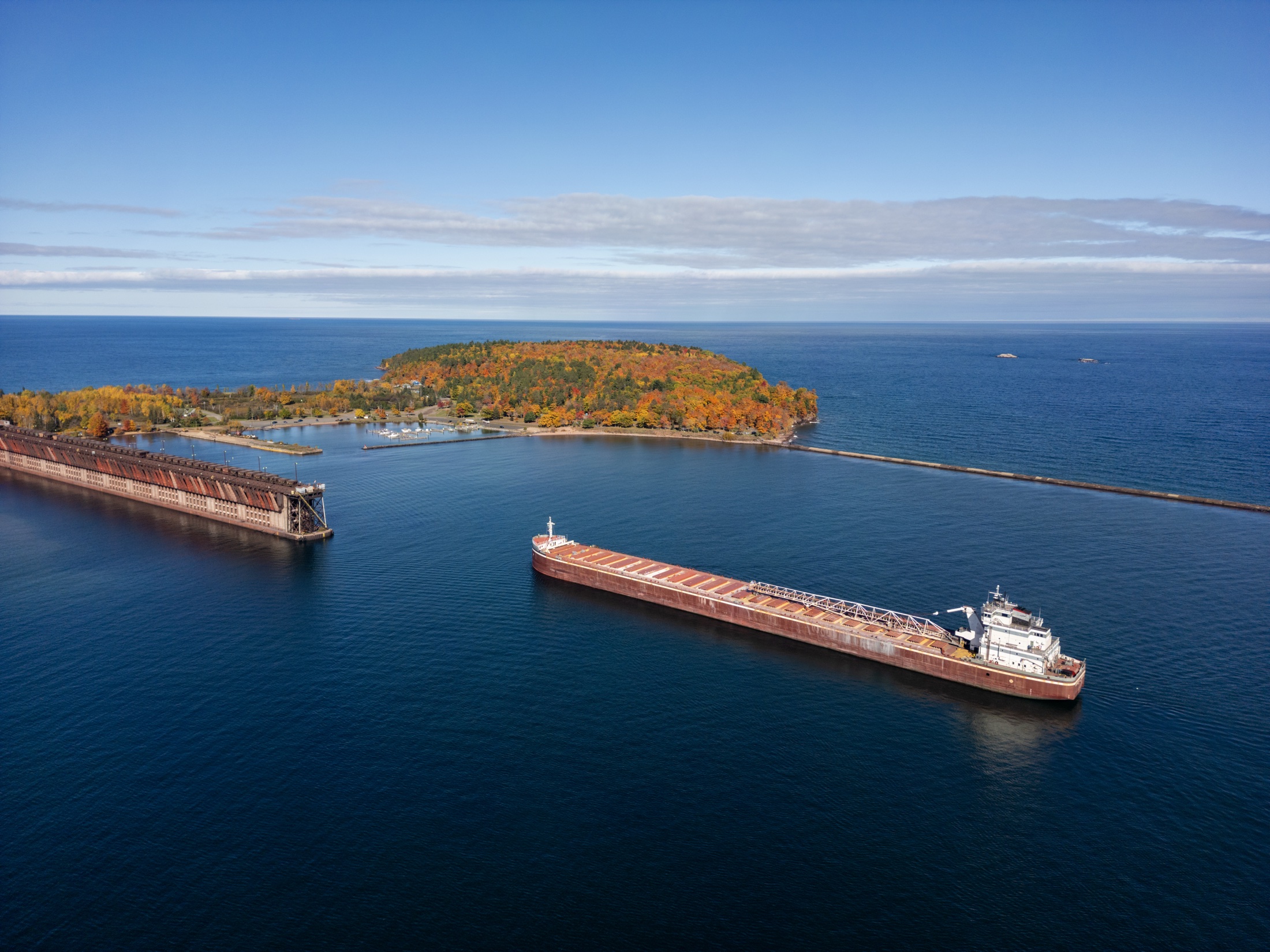 Aerial view of a harbor and rocky shoreline with a red lighthouse and blue water.