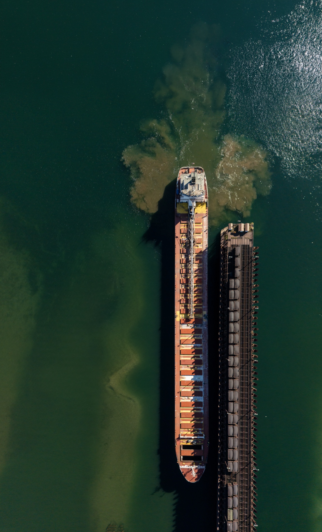 Vertical aerial view of a large ship beside an industrial dock in dark green water.