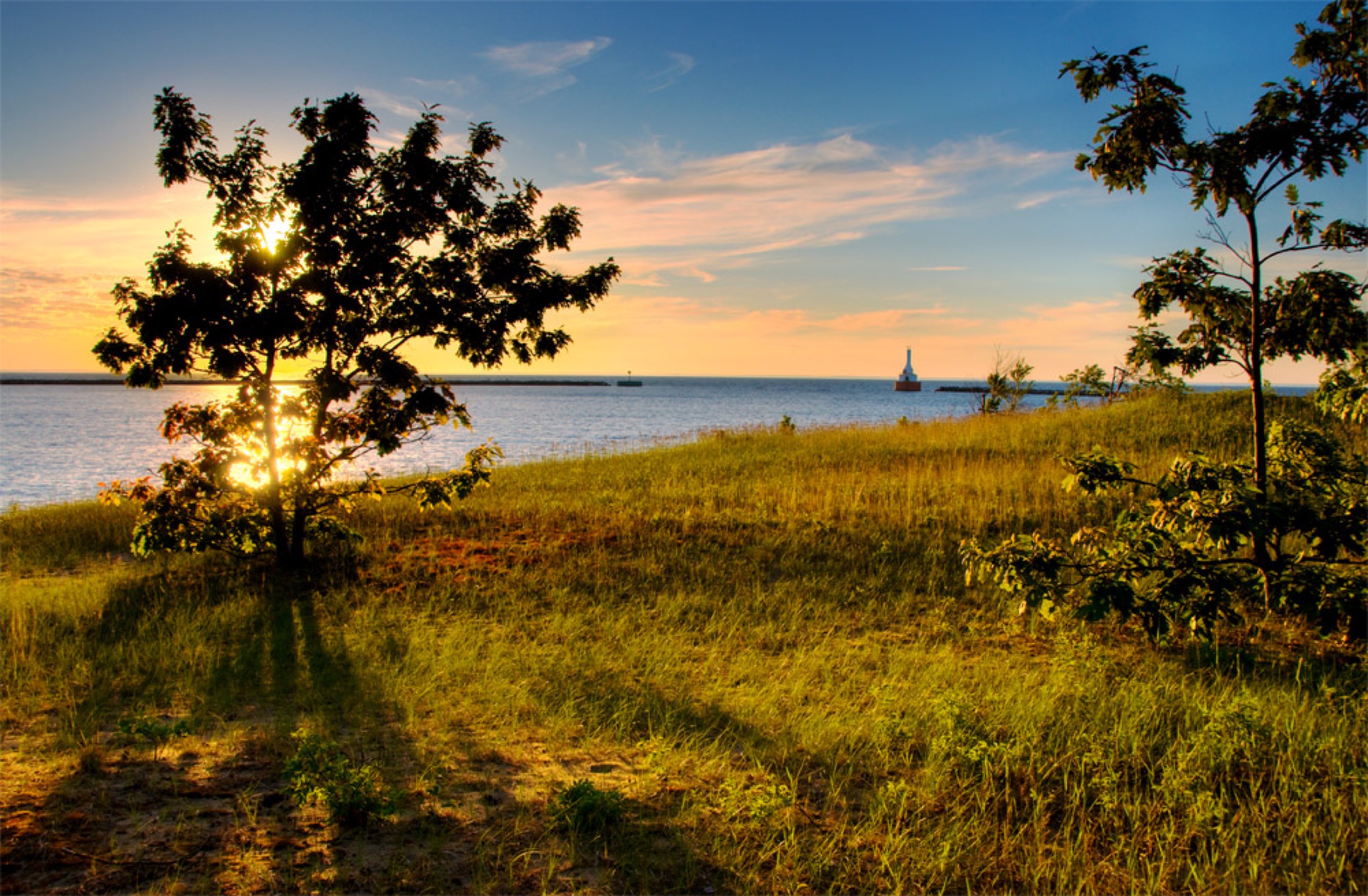 Sunset over water and grass with trees in silhouette and a small lighthouse in the distance.
