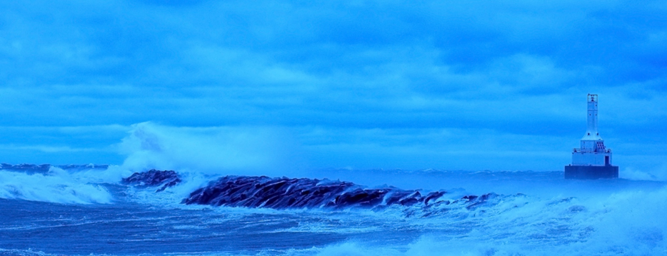 Blue storm waves breaking against rocks with a lighthouse in the distance.