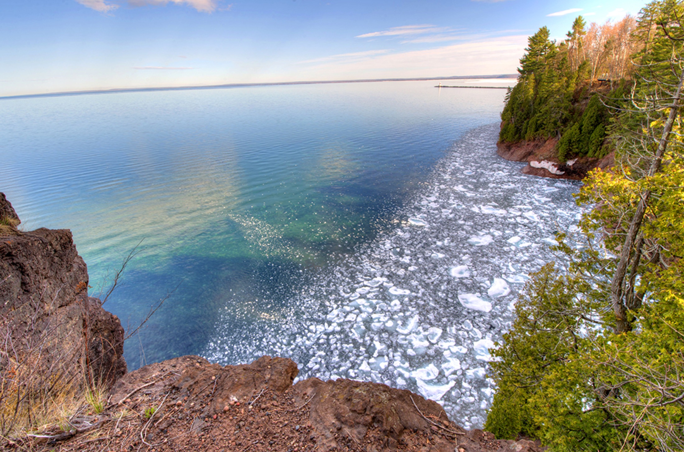 View from rocky cliffs over blue water, shoreline trees, and floating ice.