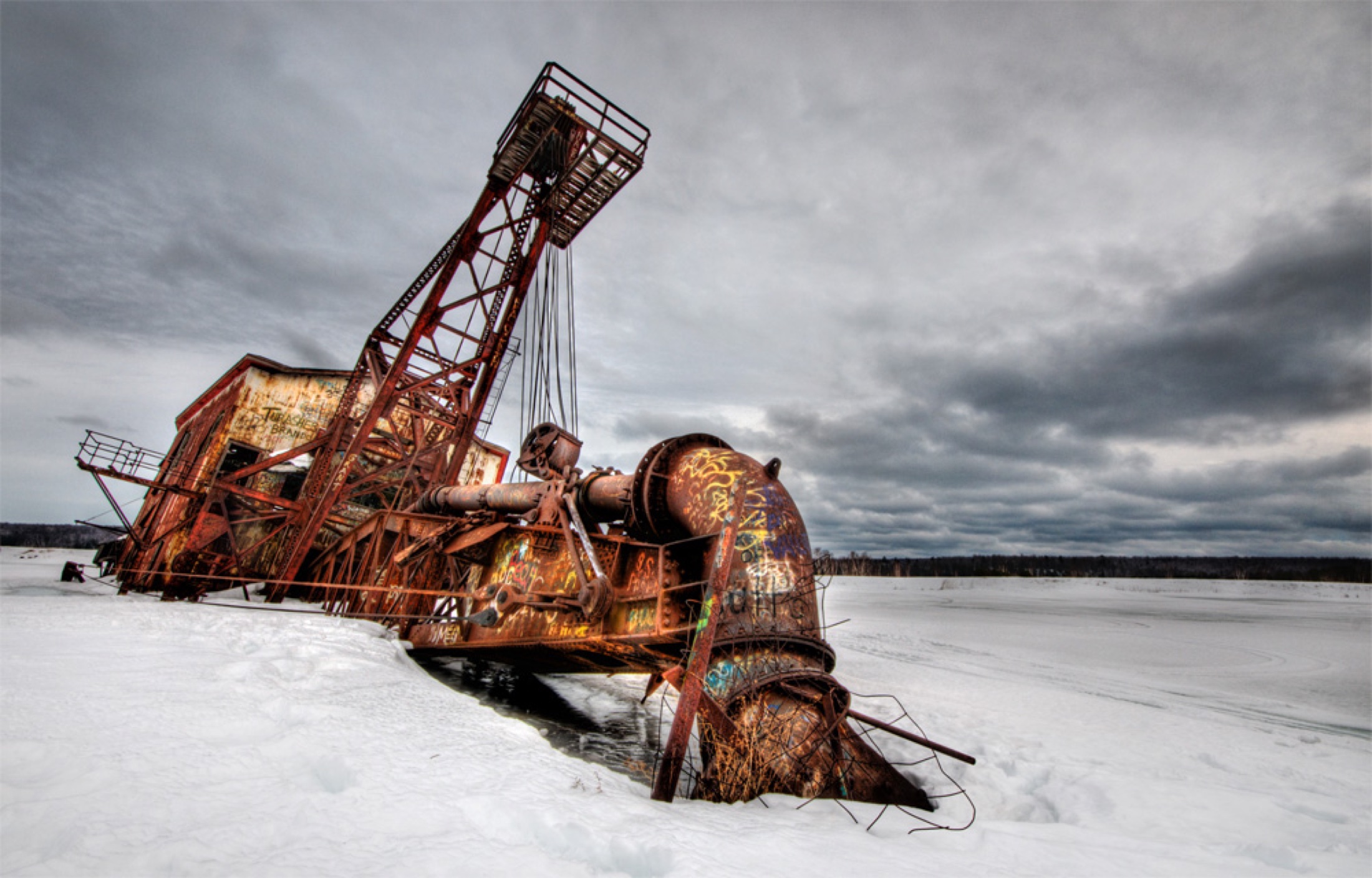 Rusting industrial dredge structure surrounded by snow under a heavy gray sky.
