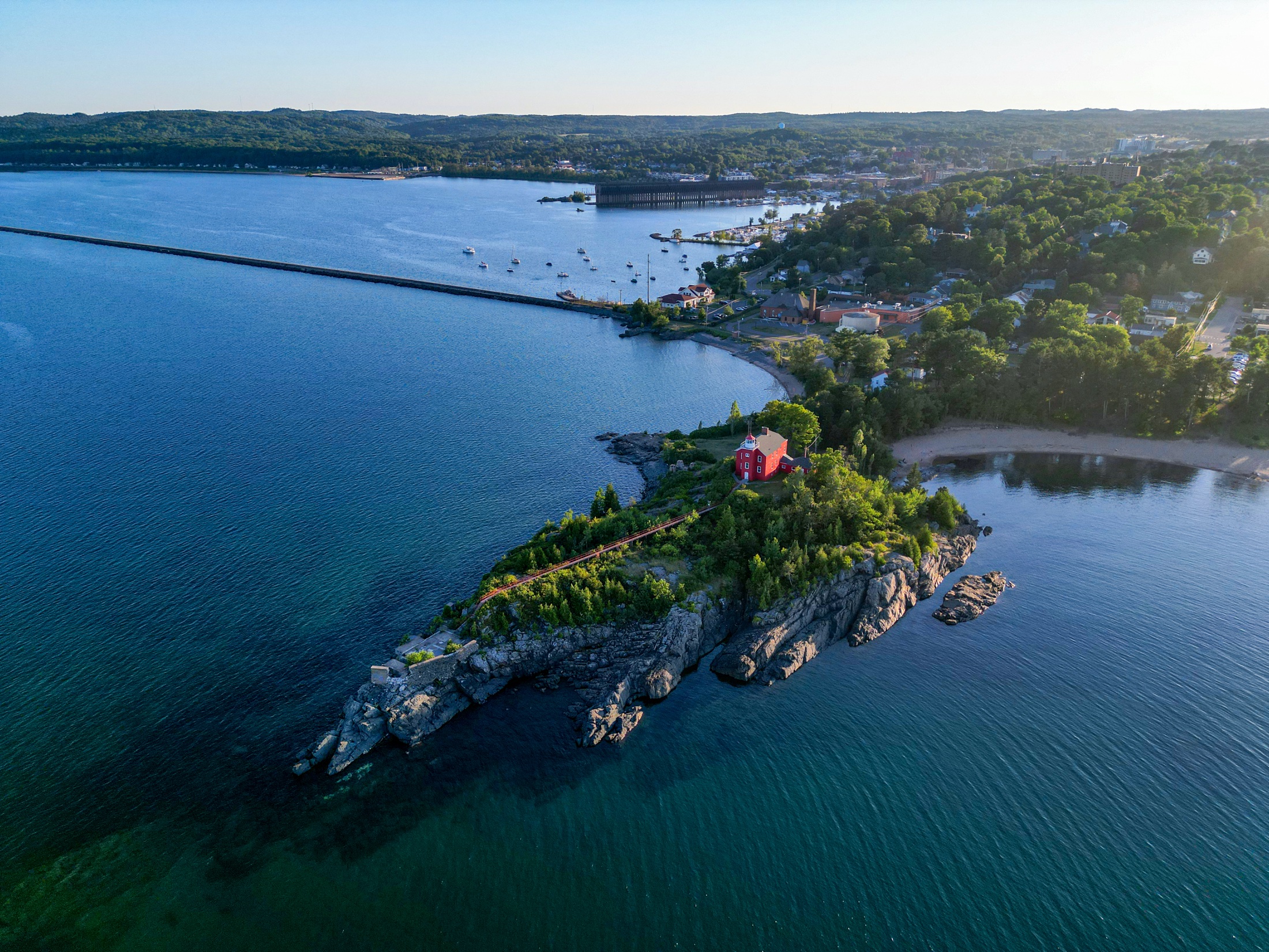 Aerial view of a red lighthouse on a rocky point surrounded by blue water.