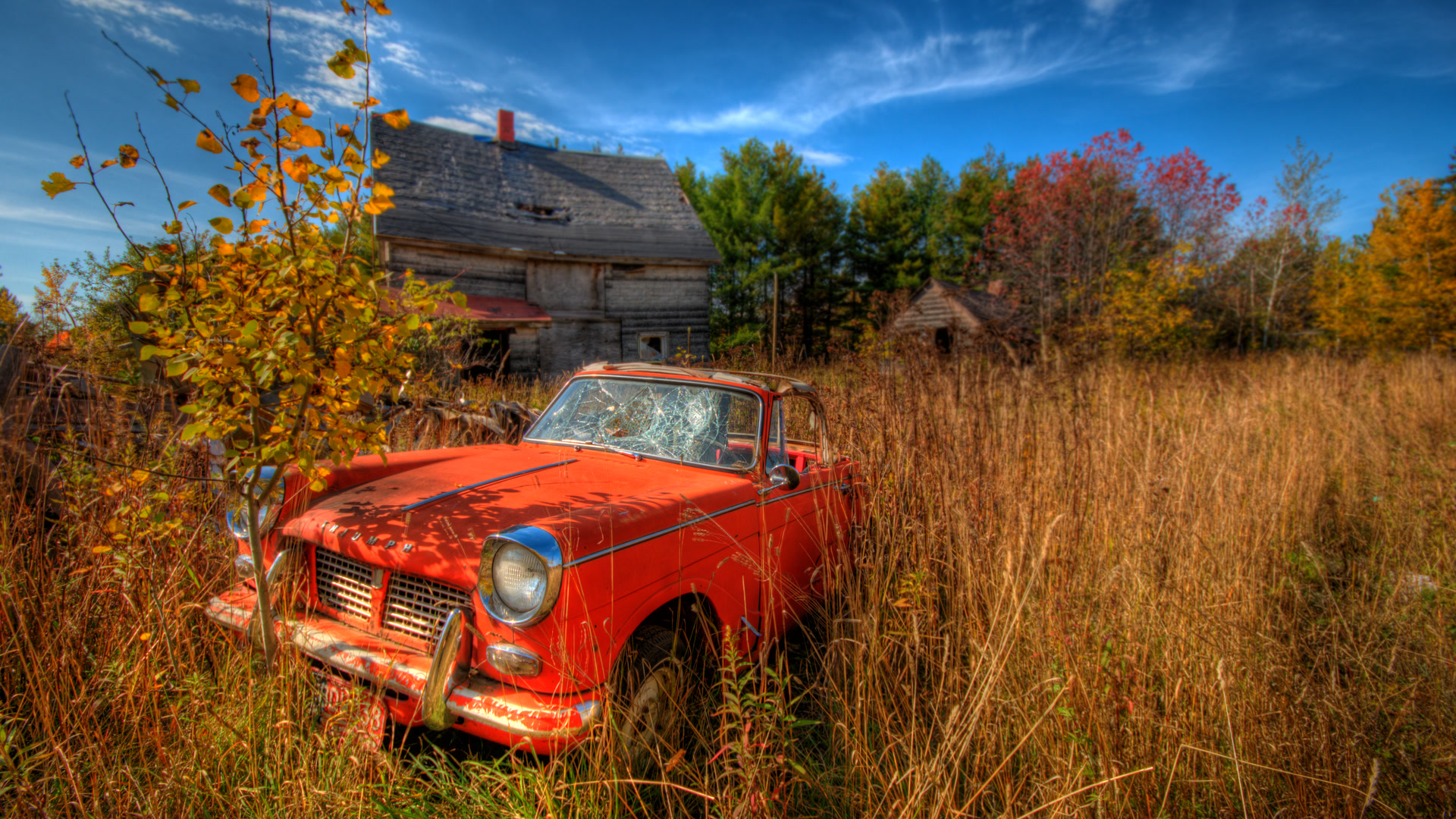 Original photograph: an abandoned red convertible in autumn grass beside weathered wooden buildings.